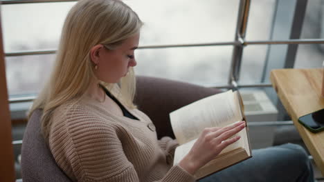 lady seated thoughtfully flipping through pages of book with phone and coffee cup on table in cozy cafe or reading area, focused on her reading experience