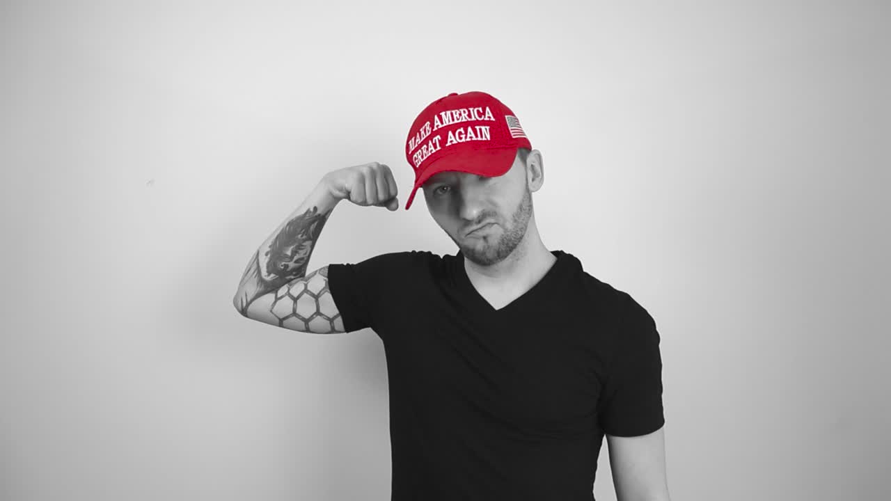 Grayscale footage of a tattooed man wearing a red MAGA Make America Great Again hat lifting up his tattooed full sleeve arm and showing his biceps muscle and seems happy with it. American patriot.
