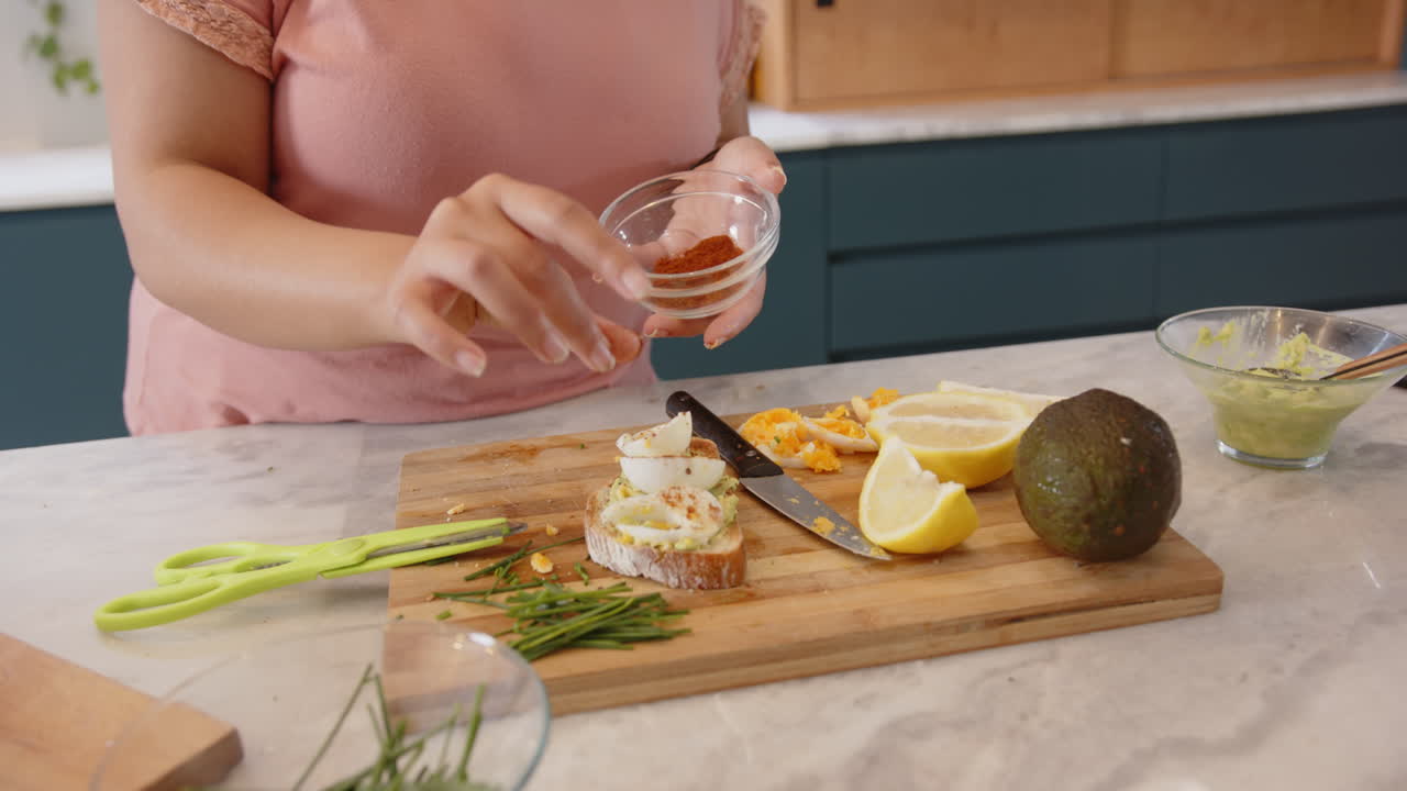 Preparando aperitivos saludables, mujer añadiendo condimento a una tostada de aguacate en la cocina.