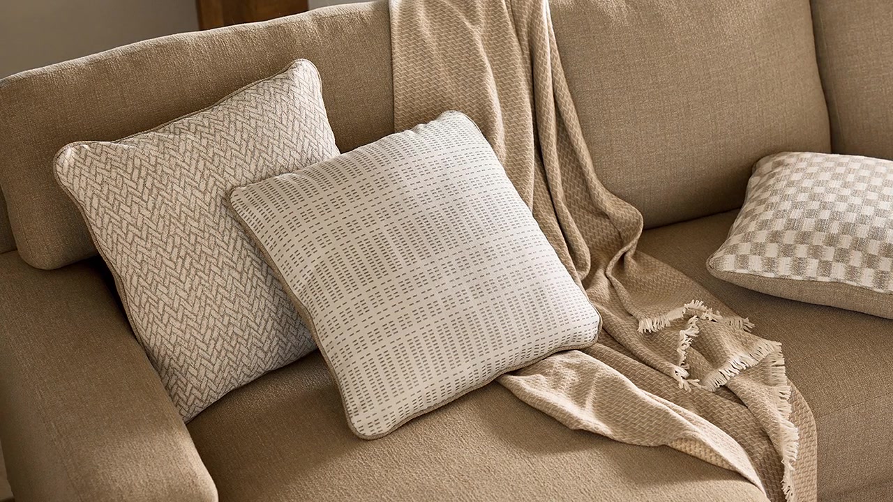 Lento zoom su due cuscini decorativi disposti su un divano in tessuto marrone con una coperta plaid in un soggiorno allestito professionalmente.