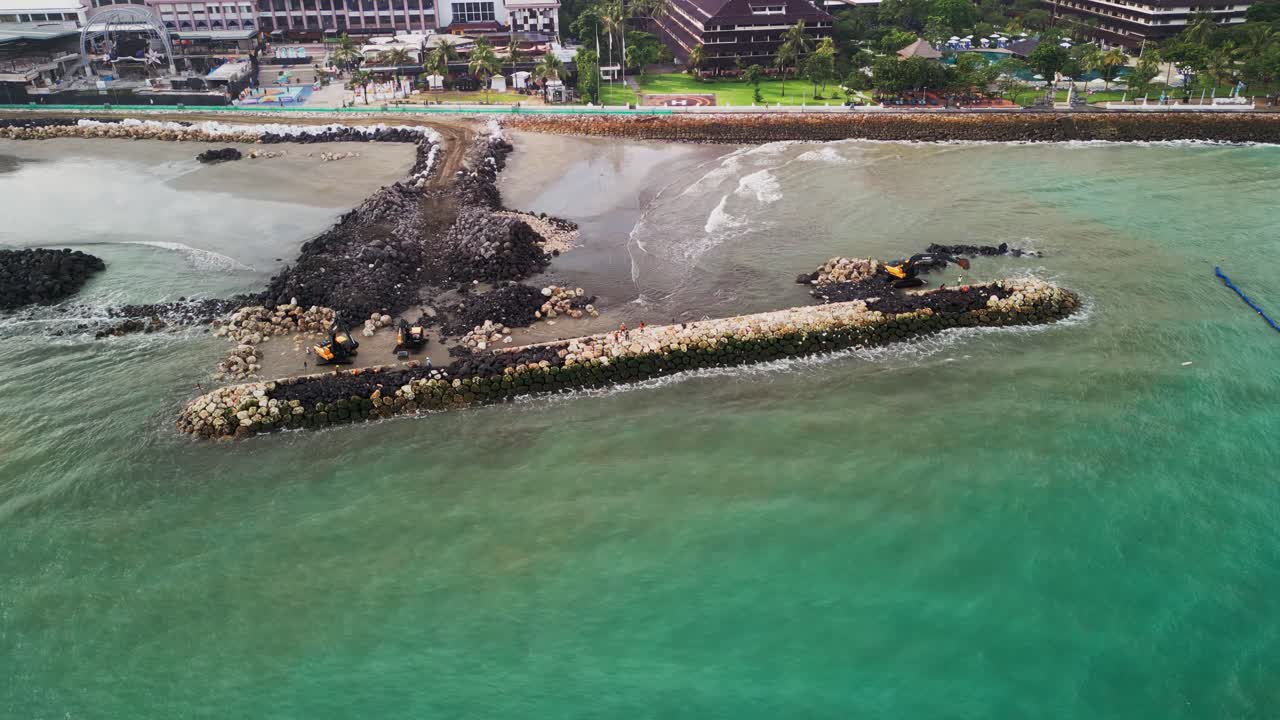 Coastal construction is underway as excavators carefully position boulders along the shoreline to form wave breakers while ocean waves hit the structure in an ongoing effort to preserve the land.