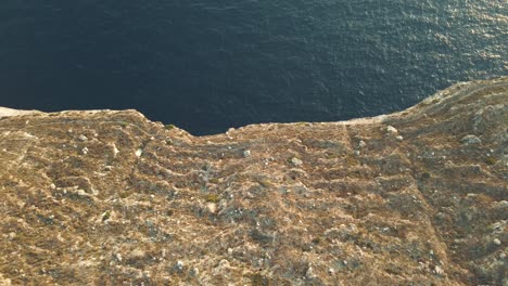 Cinematic-aerial-view-looking-down-a-vertical-cliff-over-a-peaceful-sea-slowly-opening-perspective-towards-the-horizon,-drone-footage