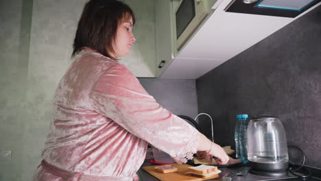 caregiver in pink velvet robe preparing multiple sandwiches with sliced bread, cheese, and cold cuts on kitchen counter near electric kettle and apples, performing morning routine in bright home setting