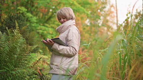 female scientist standing amid thick autumn foliage taking plant notes with tablet during ecological study, partially obscured by blurred greenery in foreground