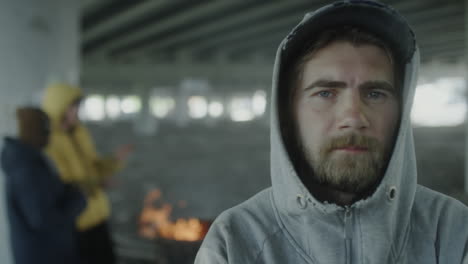 man in hoodie with beard looking at camera, people and fire in background under an overpass