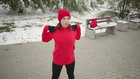 lady performing hand stretch turning her arm in outdoor fitness routine, dressed in athletic wear, background features frosted tree, bag on bench, and snow-covered ground