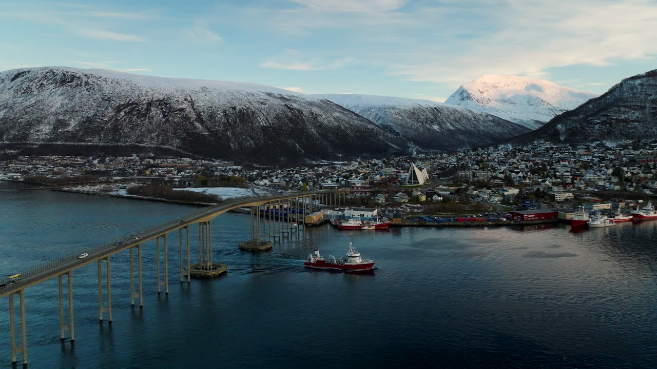 Pohled z dronu na provoz na mostě Tromsø s nákladní lodí proplouvající pod ním, město a zasněžené hory na začátku zimy, Norsko.