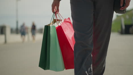 leg view of person walking on a street holding colorful shopping bags, with a hand visible carrying a handbag, blurred background features other passersby and a slightly urban outdoor