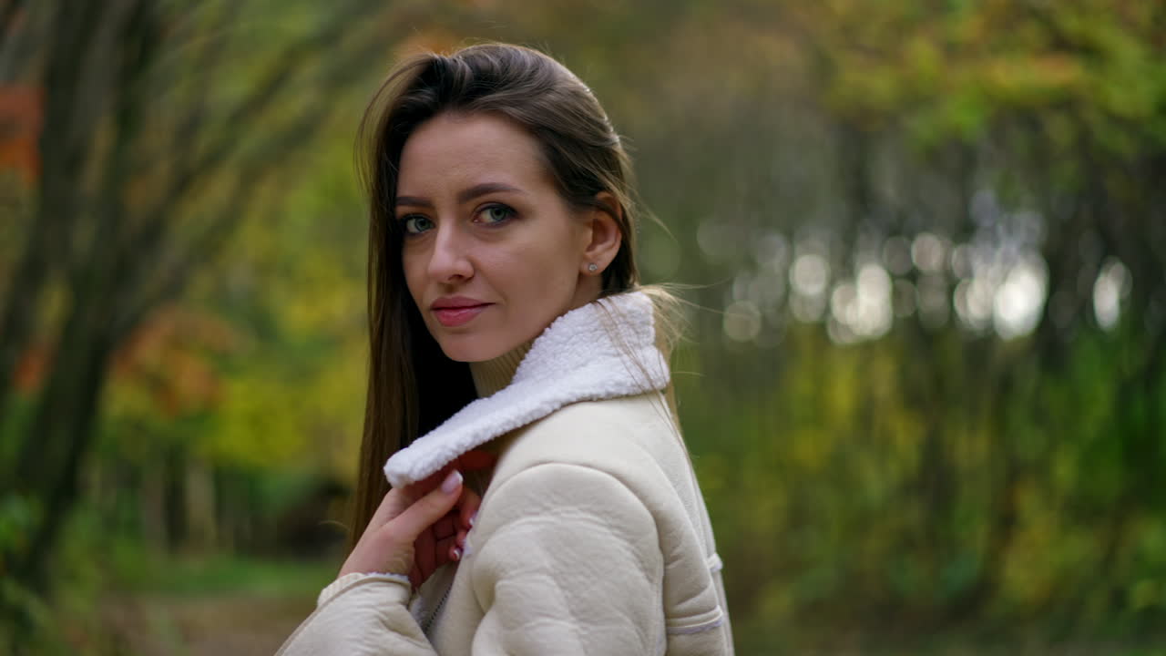 Beautiful big-eyed brunette in white jacket stands in the park her side to camera looking at it. Lady looks at the paper cup in her hands touching her hair.