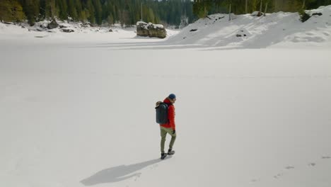 Tracking-aerial-shot-of-man-with-orange-jacket-walks-on-frozen-snow-covered-lake