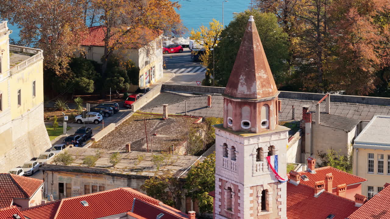 Aerial drone view of a tall church tower displaying the Croatian flag, surrounded by autumn colored trees and rooftops. Zadar, Croatia
