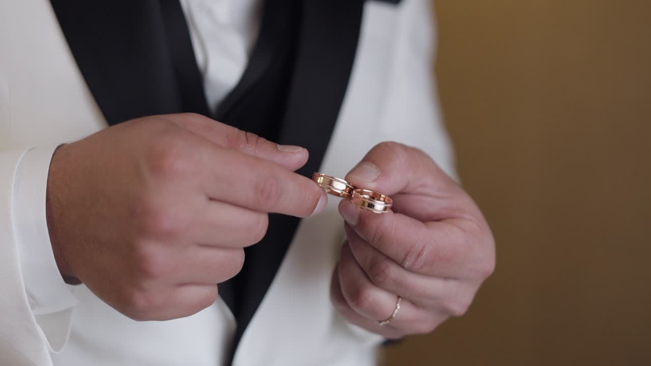 Groom in white jacket holding wedding rings on palm of his hand, man touches the rings, slow motion