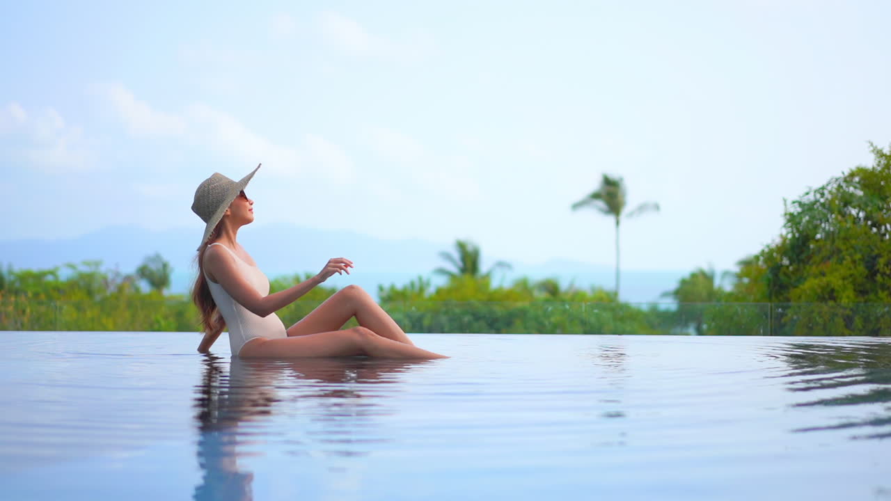 Asiatische Frau im Badeanzug und mit Hut sitzt am Rand eines Infinity-Pools, stützt sich auf einen Arm und genießt die Aussicht auf tropische Berge und grüne Bäume im Hintergrund, Zeitlupen-Seitenansicht bei Tag