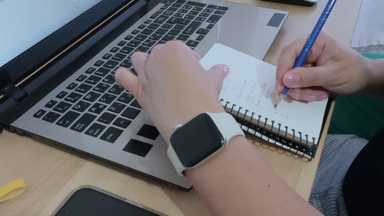 Person working remotely, writing notes in a spiral notebook while using a laptop