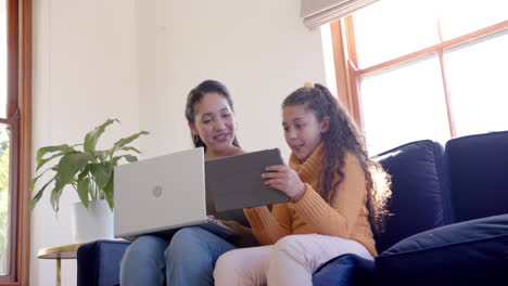 Glückliche-Biracial-Mutter-Und-Tochter-Sitzen-Auf-Dem-Sofa,-Mit-Laptop-Und-Tablet-Im-Sonnigen-Wohnzimmer