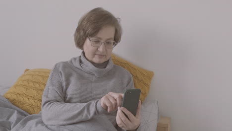 Senior-Woman-With-Eyeglasses-Sitting-On-Bed-Using-Smartphone