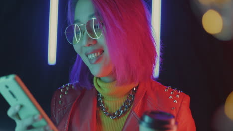stylish young woman with pink hair, drinking coffee and using smartphone at night