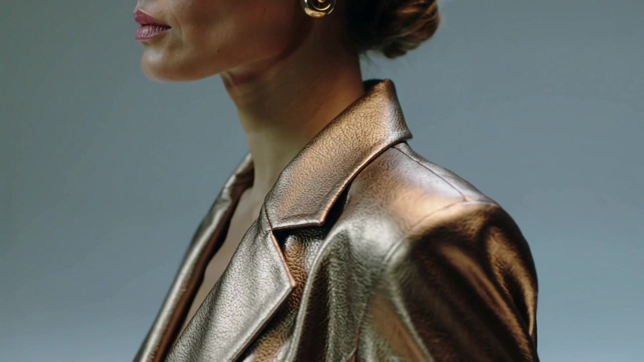 Moving camera, model in bronze blazer rotating to camera in studio, showing gold hoops and lipstick