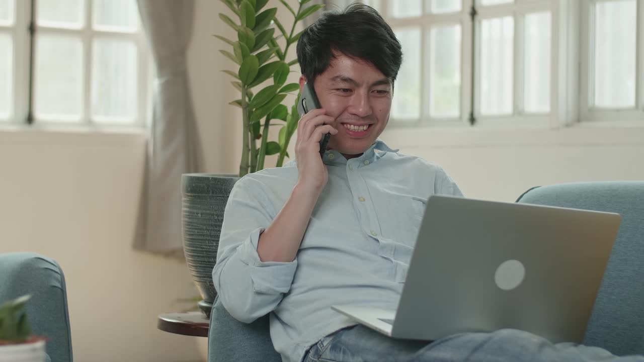 Man Sitting Sofa In Living Room While Speaking By Phone And Working On Laptop, Video In 4K