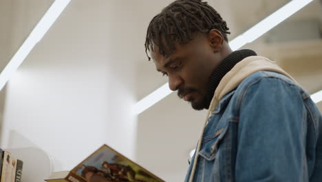 male in denim jacket studying book closely in modern library. focused young man examines pages carefully while surrounded by well-organized shelves of colorful books in peaceful library environment