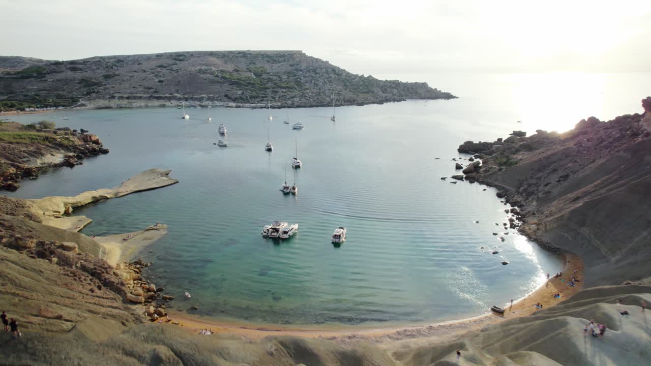 Drone moves forward over the sandy hills of Golden Bay, Malta, showing walking paths, boats on calm water, and sunlight reflection