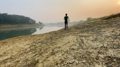 Man-jogging-in-slow-motion-by-dry-river-bed-in-Sylhet,-rear-view,-hazy-morning