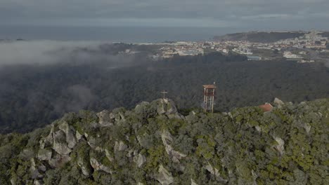 areal drone footage of a lookout tower and a building on the top of a mountain surrounded by a forest, near nazare, portugal, tourist attraction