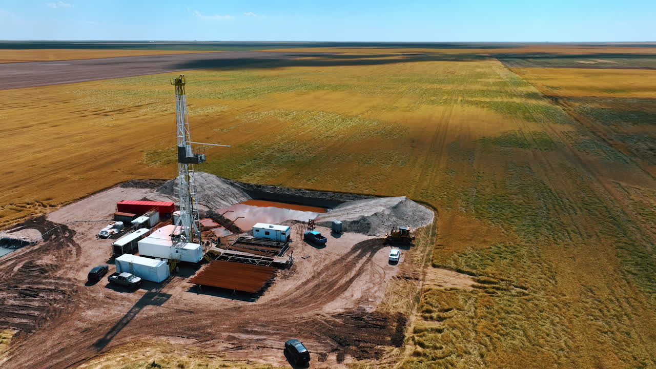 Approaching the oil production site in the immense field. Derrick in the center of location, equipment for drilling resources, two water pools and cars around the rig.