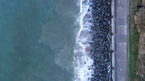 Toma-Aérea-Recta-Hacia-Abajo-De-Un-Paseo-De-Hormigón-Con-Olas-Golpeando-Las-Defensas-Marítimas