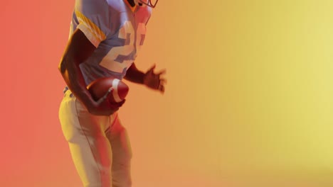 Video-of-african-american-american-football-player-with-ball-over-yellow-to-orange-background