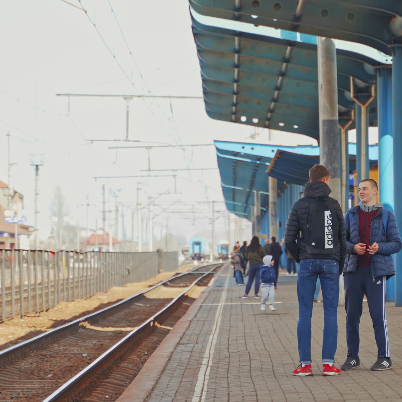 Two teenagers standing on a railway platform waiting for a train. Young men in sportive clothes talking to each other on a railway station outdoors.