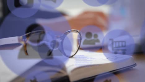 animation of technology icons over book, glasses and hands of caucasian man typing on keyboard