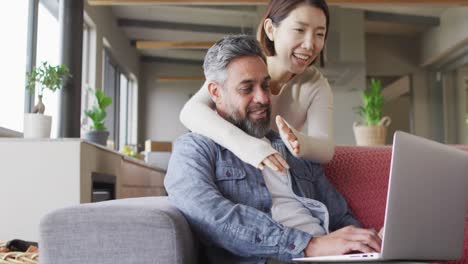 una pareja feliz y diversa sentada en la sala de estar usando una computadora portátil y hablando