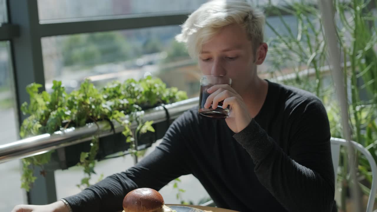 은 머리카락의 젊은이가 도시 전망, 자연 빛, 비 식물과 함께 창 에 있는 카페 테이블에서 샌드위치와 얼음 커피를 즐기고 있습니다. 편안하고 캐주얼한 식사 장면