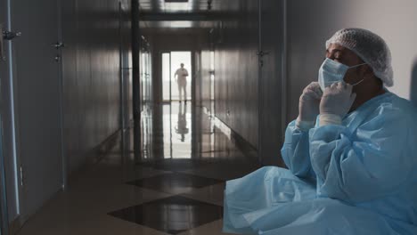 tired doctor in blue gown, medical cap and gloves sitting in the hallway while removing his mask, in the background another doctor walks