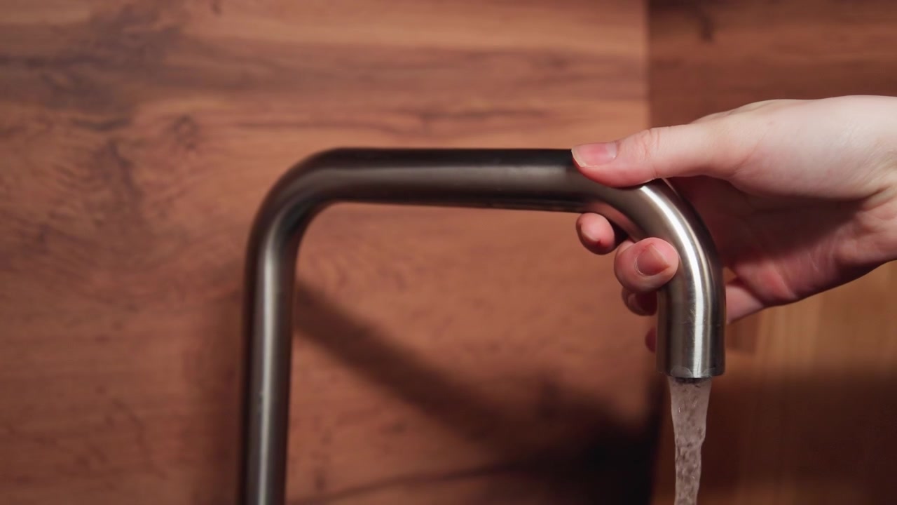 Brushed metal faucet pours clear water into sink near warm wood backsplash, with reflective highlights and gentle flowing movement suggesting clean kitchen use, home routine and practical plumbing