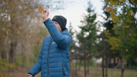 russian kid in blue jacket and beanie bends down to pick yellow leaf from ground, observes it carefully before placing it in paper bag while standing in vibrant autumn outdoor setting