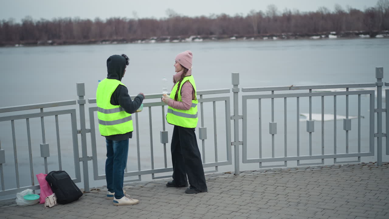 Two people in reflective safety vests standing on riverside promenade near metal railing, talking and taking break, holding food and drinks, bags placed on ground, cold overcast day