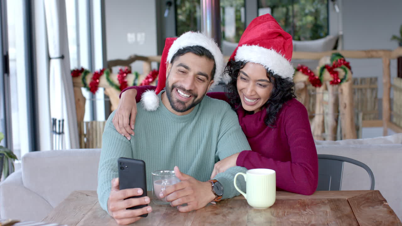 una pareja biracial feliz con sombreros de santa claus usando un teléfono inteligente para una llamada de video en casa, en cámara lenta