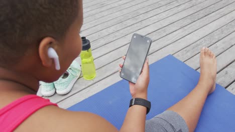 happy african american woman practicing yoga, sitting using smartphone on deck in garden