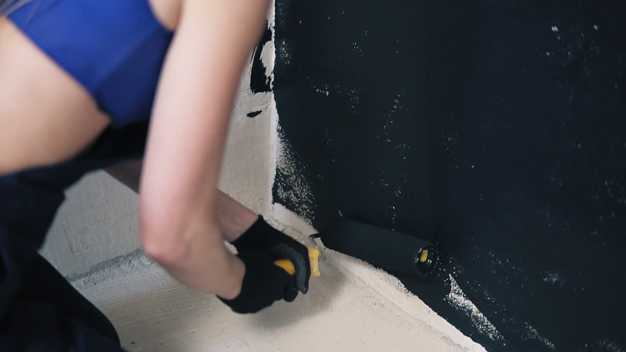 Young woman with paint roller carefully paints the wall with black colour