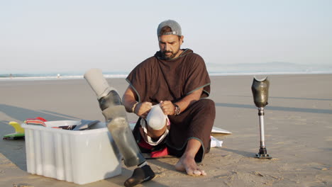 front view of a pensive male surfer sitting on the beach and putting prosthetic leg on