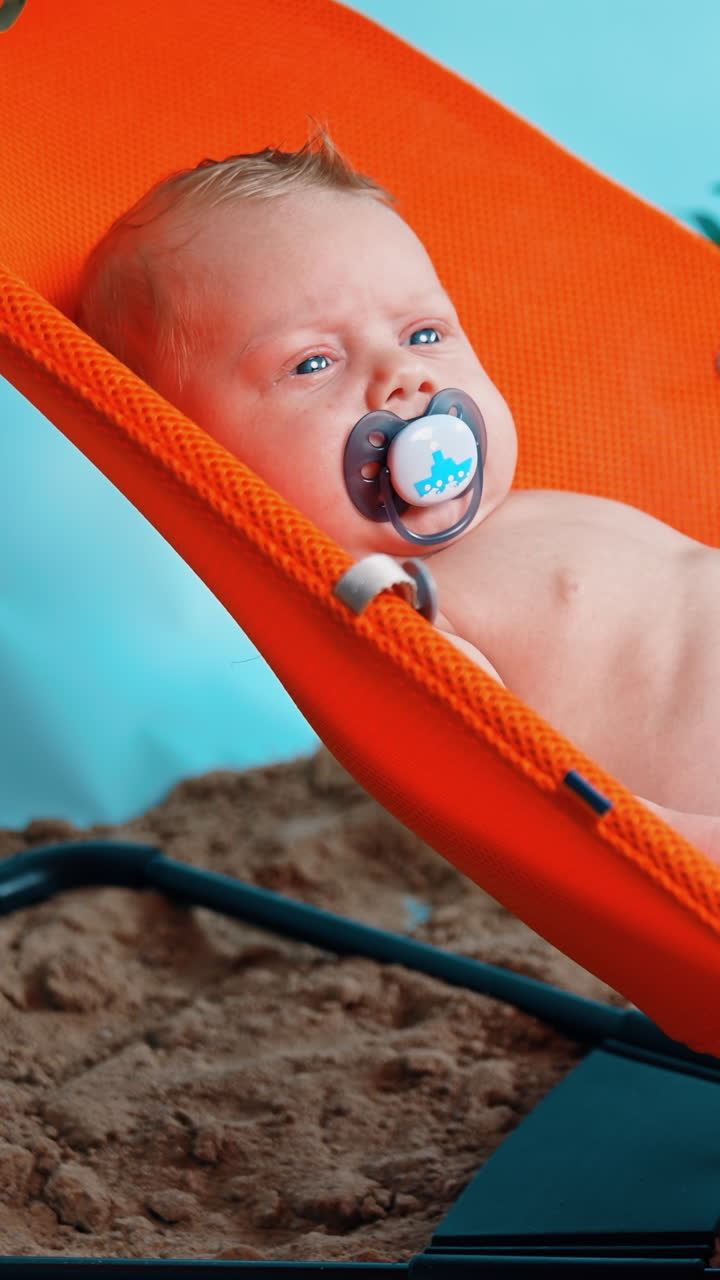 Lovely baby boy with pacifier in mouth lies in chair. Infant is winking from the bright light. Vertical video