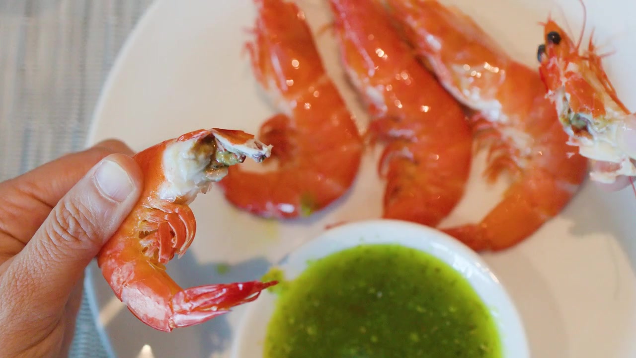 Primo piano di mani che sbucciano gamberi grigliati e li intingono in salsa piccante di peperoncino verde.