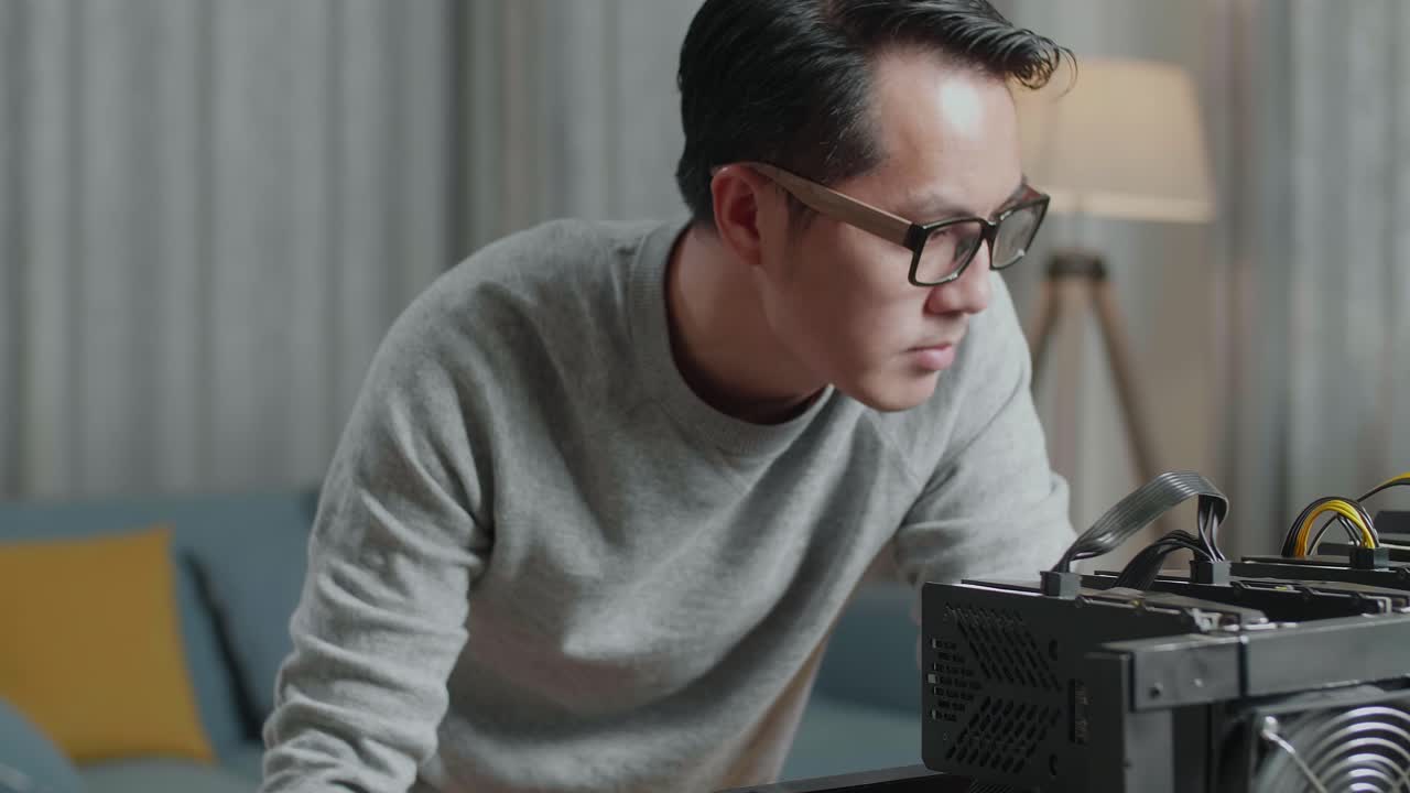 안경을 쓴 만족스러운 아시아 남자가 노트북으로 일하면서 암호화폐 채굴을 위한 채굴 기계를 보고 있다.
