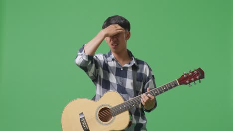 asian teenager with guitar having a headache while standing on green screen background