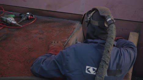 Welder-on-shipyard-taking-off-mask-for-a-second-to-take-a-better-look-at-his-work,-then-putting-it-back-on-and-crawling-into-a-tight-spot