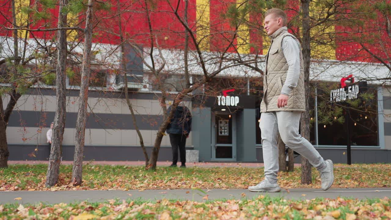 Man in jogger and sneakers walking as breeze blows dry foliage around, trees sway gently, with colorful building in background showing inscription, and several people nearby in lively urban setting