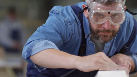 man in safety goggles sanding timber with sandpaper and then blowing dust away