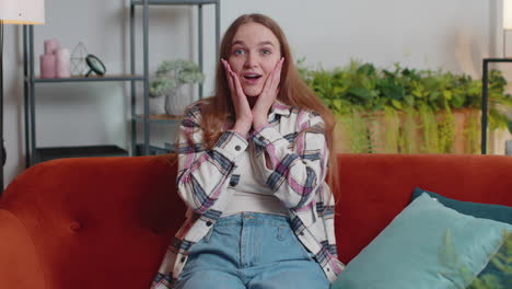 excited amazed young woman raising hands in surprise shocked by sudden victory at home on sofa